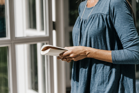 Frau mit Buch am Fenster stehend, Teilansicht, lizenzfreies Stockfoto