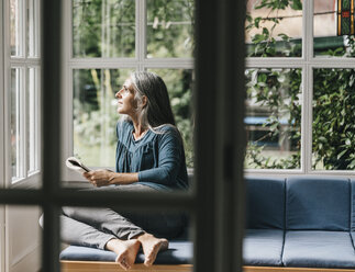 Entspannte Frau sitzt mit einem Buch auf dem Sofa im Wintergarten und schaut durch das Fenster - KNSF000316