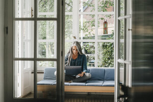 Woman sitting on lounge in winter garden using laptop - KNSF000324