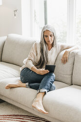 Frau mit langen grauen Haaren sitzt auf der Couch mit E-Book - KNSF000343