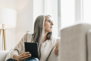 Frau mit E-Book sitzt auf der Couch und schaut durch das Fenster - KNSF000344