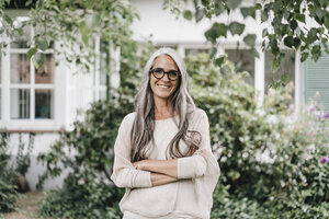 Portrait of smiling woman with long grey hair wearing spectacles standing in the garden - KNSF000356