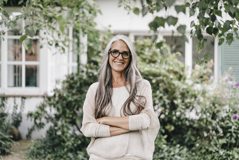 Porträt einer lächelnden Frau mit langen grauen Haaren und Brille, die im Garten steht - KNSF000356