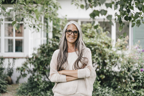 Portrait of smiling woman with long grey hair wearing spectacles standing in the garden - KNSF000356