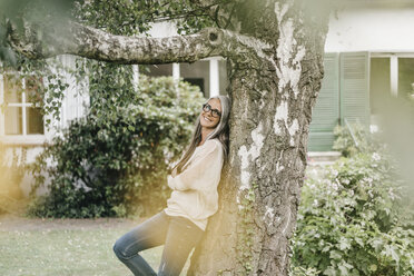 Lächelnde Frau lehnt an einem Baum im Garten - KNSF000358