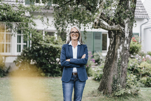 Porträt einer lächelnden Geschäftsfrau mit langen grauen Haaren und Brille, die im Garten steht - KNSF000361