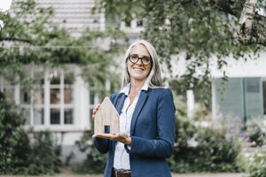 Porträt einer lächelnden Geschäftsfrau mit einem Stück Holz in Form eines Hauses, das im Garten steht - KNSF000363