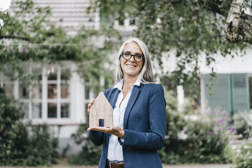 Portrait of smiling businesswoman with piece of wood shaped like a house standing in the garden - KNSF000363