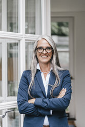 Portrait of smiling businesswoman with long grey hair and spectacles standing in front of open glass door - KNSF000367