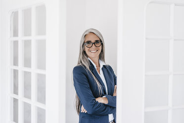 Portrait of smiling businesswoman with long grey hair and spectacles standing between two glass doors - KNSF000368