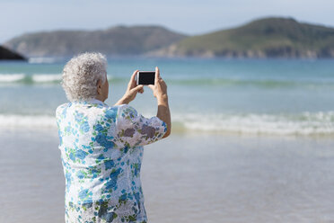 Rückansicht einer älteren Frau am Strand, die ein Foto mit ihrem Smartphone macht - RAEF001427