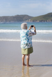 Rückansicht einer älteren Frau am Strand, die ein Foto mit ihrem Smartphone macht - RAEF001428