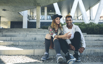 Lächelndes junges Paar sitzt auf einer Treppe und schaut auf ein Smartphone - DAPF000274