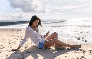 Spain, Asturias, beautiful young woman with smartphone on the beach - MGOF002209