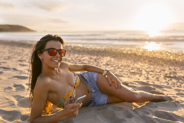 Spanien, Asturien, schöne junge Frau mit Smartphone am Strand bei Sonnenuntergang - MGOF002238