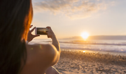 Spain, Asturias, beautiful young woman taking a picture on the beach at sunset, rear view - MGOF002239