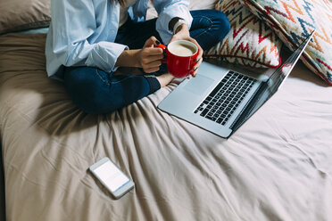 Young woman sitting on bed, coffee cup, laptop and smartphone - BOYF000569