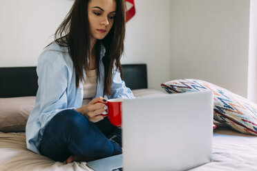 Young woman sitting on bed, coffee cup, laptop and smartphone - BOYF000573