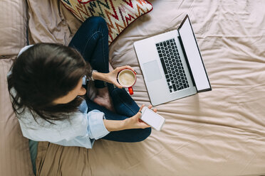 Junge Frau sitzt auf dem Bett, Kaffeetasse, Laptop und Smartphone - BOYF000576