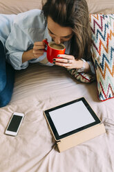 Junge Frau auf dem Bett sitzend, Kaffeetasse, digitales Tablet und Smartphone - BOYF000580