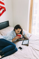 Junge Frau auf dem Bett sitzend, Kaffeetasse, digitales Tablet und Smartphone - BOYF000581