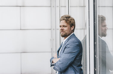 Confident businessman standing at glass front of a building - KNSF000384