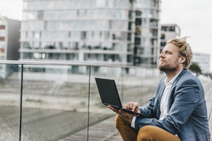 Businessman sitting on bridge using laptop looking up - KNSF000394