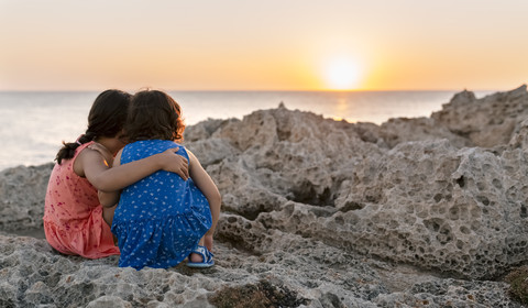 Back view of two little sisters crouching at rocky coast watching sunset stock photo