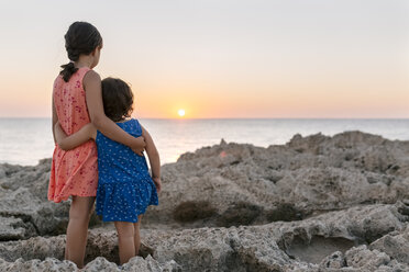 Back view of two little sisters standing arm in arm at rocky coast watching sunset - MGOF002254