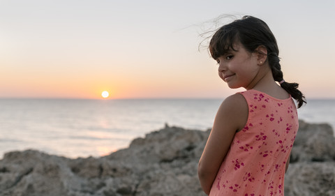 Lächelndes kleines Mädchen an felsiger Küste bei Sonnenuntergang, lizenzfreies Stockfoto