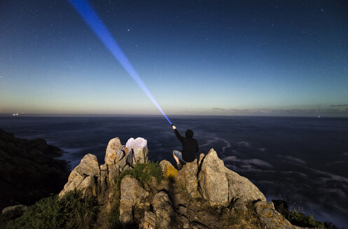 Mann sitzt auf Felsen an der Küste und leuchtet mit einer Taschenlampe in den Himmel - RAEF001437