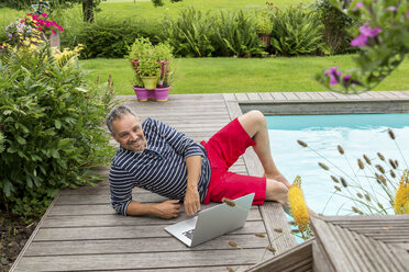 Smiling man sitting at pool edge with his laptop - JUNF000594