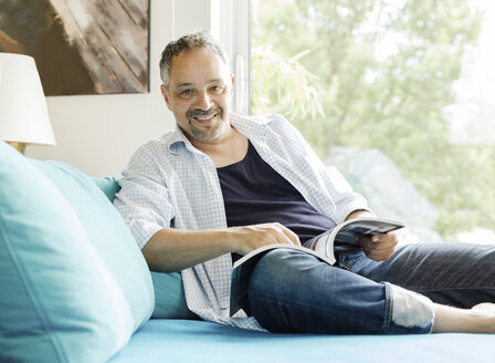 Porträt eines lächelnden Mannes, der mit einer Zeitschrift auf der Couch sitzt - JUNF000597
