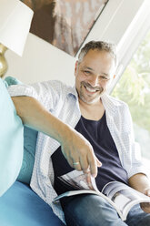 Porträt eines lächelnden Mannes, der mit einer Zeitschrift auf der Couch sitzt - JUNF000598