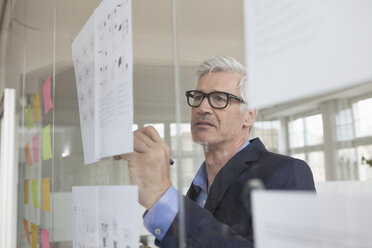 Businessman in office writing on paper at glass pane - RBF005035