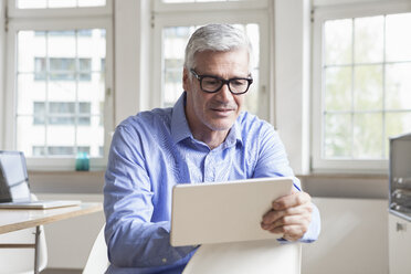 Mature businessman sitting on chair in office holding tablet - RBF005043