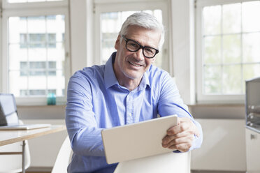 Portrait of smiling mature businessman sitting on chair in office holding tablet - RBF005044