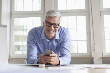 Porträt eines lächelnden reifen Geschäftsmannes am Bürotisch, der ein Mobiltelefon hält - RBF005048