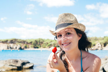 Portrait of smiling woman wearing straw hat on the beach holding strawberry - GEMF000979