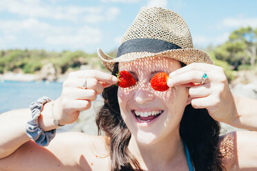 Porträt einer lächelnden Frau mit Strohhut am Strand, die ihre Augen mit Erdbeeren bedeckt - GEMF000982