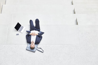 Businesssman lying on stairs next to laptop and cell phone - FMKF002936