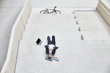 Businesssman lying on stairs next to laptop - FMKF002937