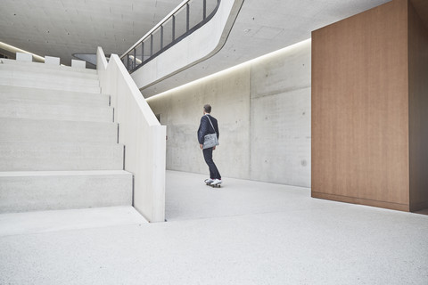 Businesssman riding skateboard along concrete wall in office building stock photo