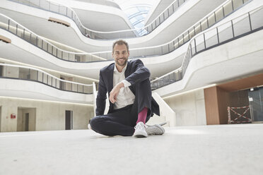 Lächelnder Geschäftsmann sitzt auf dem Boden in einem Bürogebäude - FMKF002951