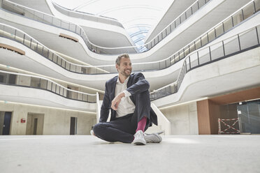 Smiling businessman sitting on the floor in office building - FMKF002952