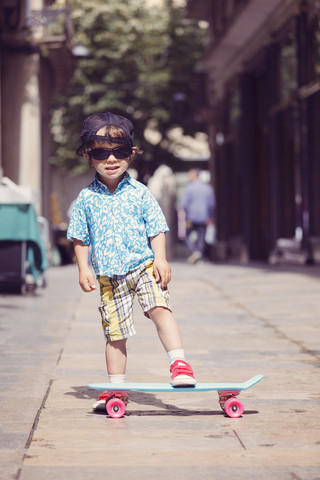 Porträt eines kleinen Jungen mit Skateboard und übergroßer Sonnenbrille und Basecap, lizenzfreies Stockfoto