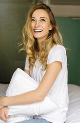 Lachende blonde Frau sitzt auf einem Bett und hält ein Kissen - JUNF000615