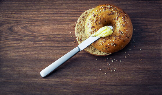 Aufgeschnittener Bagel mit Samen, Butter und einem Messer auf dunklem Holz - PPXF000032