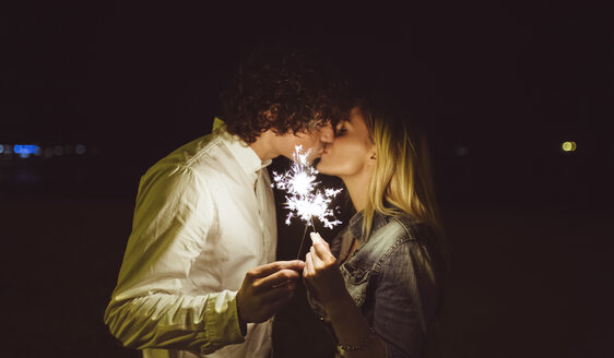Kissing young couple holding sparklers on the beach at night - DAPF000300
