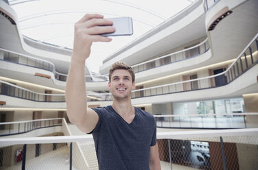 Lächelnder junger Mann macht ein Selfie in einem modernen Bürogebäude - FMKF002982